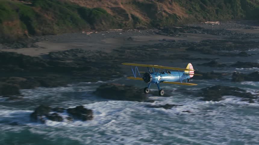 A person flies a biplane just off the coast of a landmass during the day