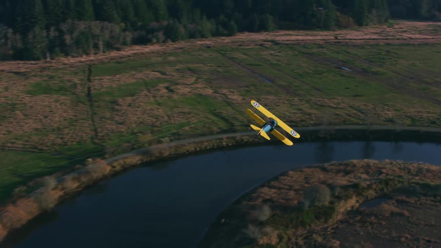 Colorful biplane flying over the river crossing the farmland at morning