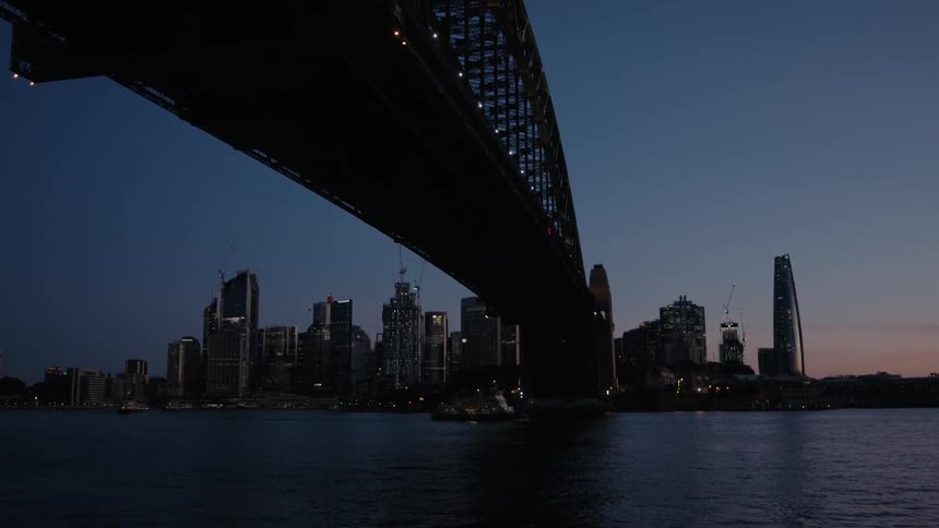A ship passes under a bridge with a city skyline in silhouette on the horizon