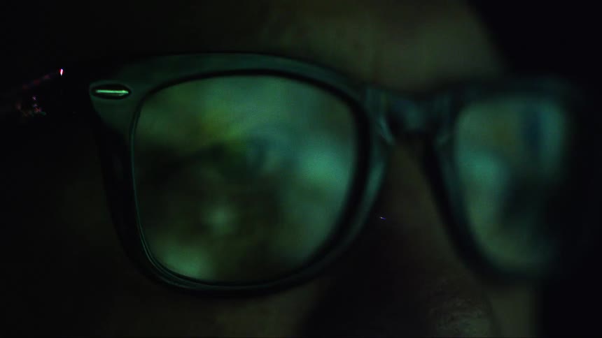 Close up of colorful lights reflecting on the glasses on the face of a man inside a room