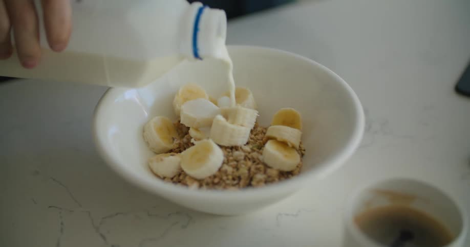 A person pouring milk on a bowl filled with cereal and banana slices for breakfast