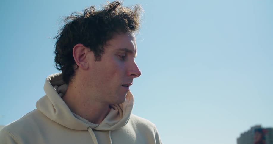 A man with a hoodie sitting and using a laptop with buildings the sky and clouds in the background
