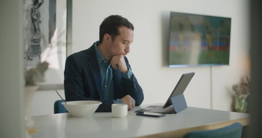 A man sits at a table at home and takes a drink out of a mug and then uses his laptop while the television plays in the background