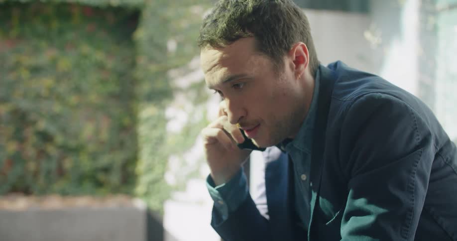 A man sits in front of a laptop talking on the phone and takes a drink from a small mug