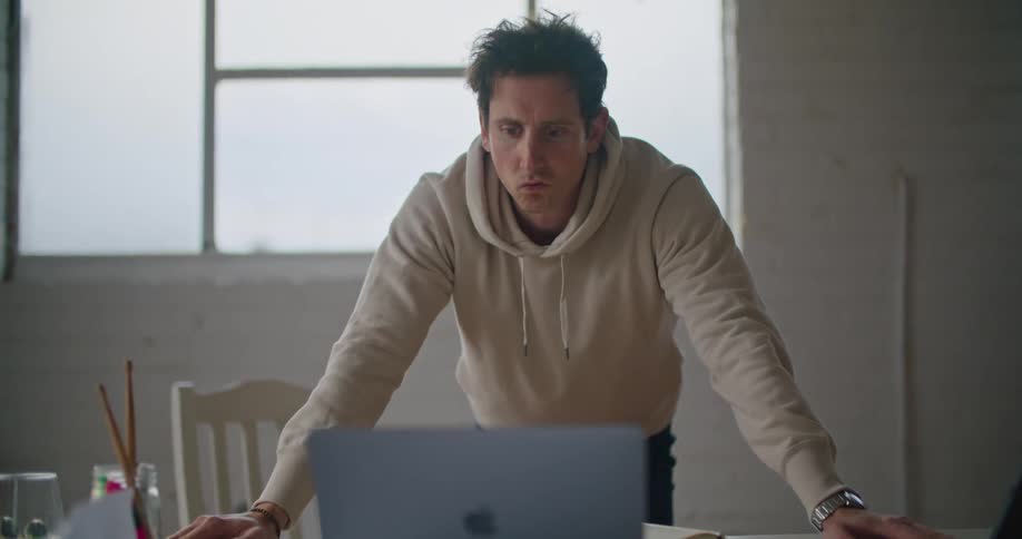 A man leaning against a table is looking at a laptop while thinking indoors during the day