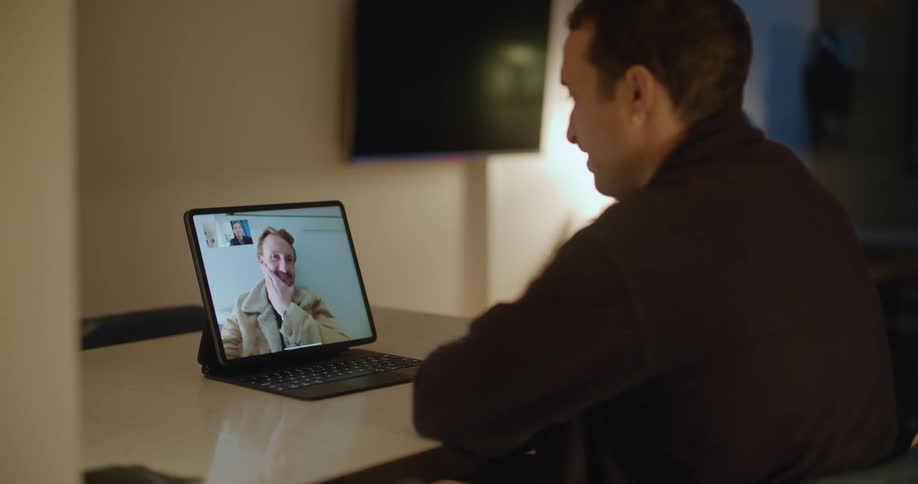 A man sits at a table with a tablet computer having a video call with another man