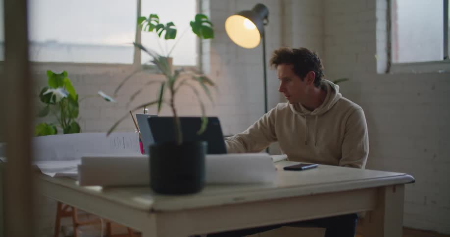 Man sits at white table consulting notebook while video conferencing over a laptop