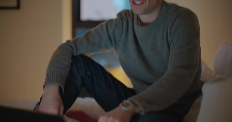 A man smiles and laughs as he talks on a video call on a laptop while sitting on a bed