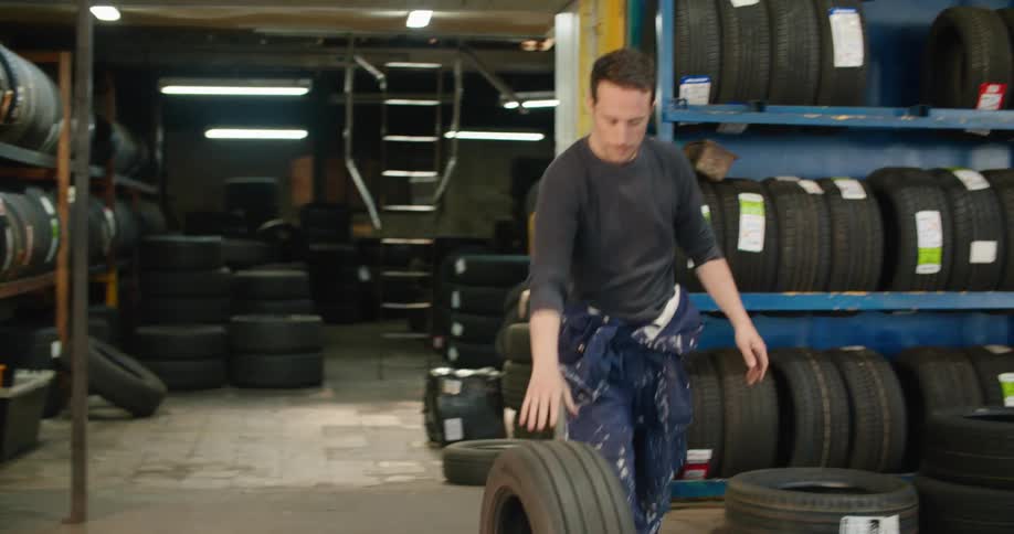A man rolls a tire across the floor then lifts it up