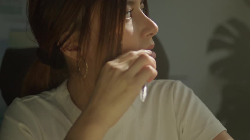 A teenage girl looks around as she chews on a pen and thinks and then writes in a book