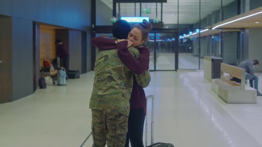 A man with military fatigues and a woman hugging in a terminal with a door and signs in the background