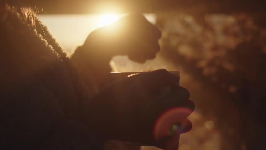 Woman wearing a fuzzy robe is backlit by bright sun as she dips tea bag into cup