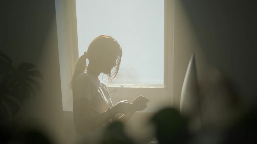 A woman stands in front of a sunny window and looks at her phone