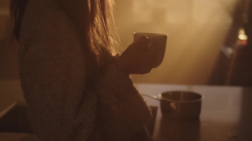 A woman holding a coffee cup while standing in a home kitchen gazes out of a window on the other side of the room