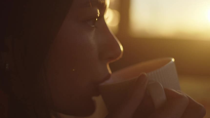 A woman sipping a cup of coffee indoors at sunrise