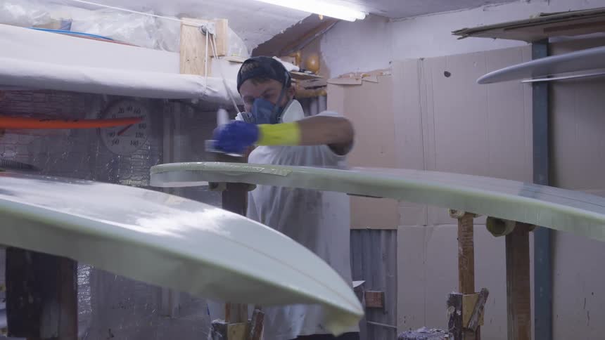 A man with a hat and mask in a room painting a surfboard with a paintbrush