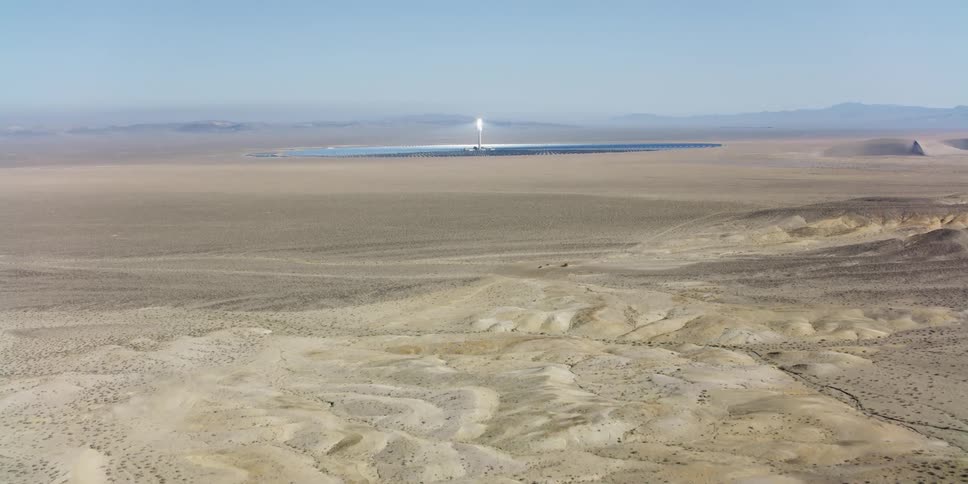 Shining tower top stands in center of facility in middle of dessert backed by misty overcast mountains on horizon