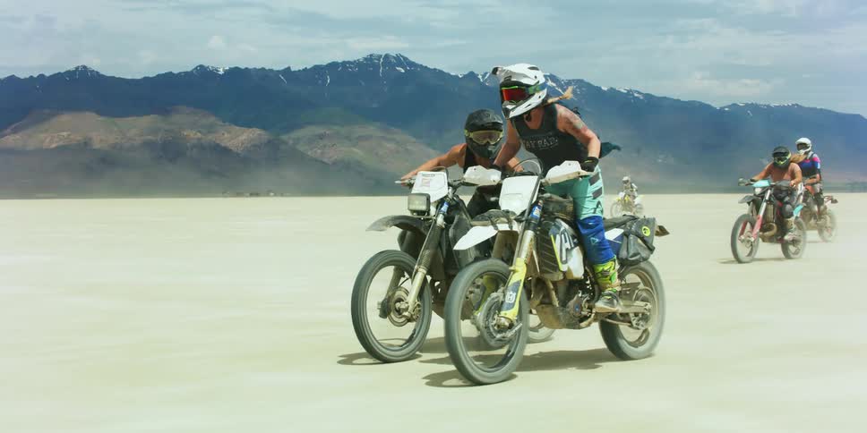 People on dirt bikes riding on a sandy desert with a mountain range sky and clouds in the background