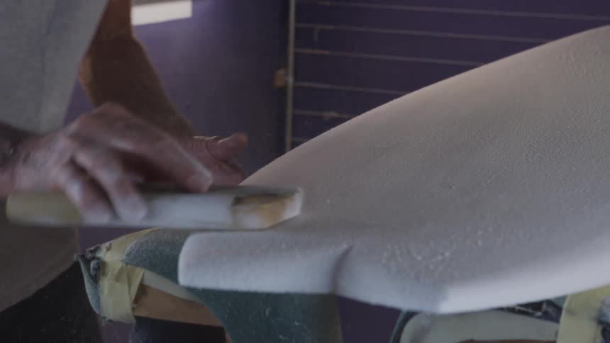 A man in a room sanding a surfboard with sandpaper with a wall in the background