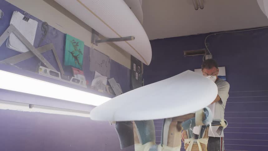 A man in a surfboard workshop examines an unfinished surfboard