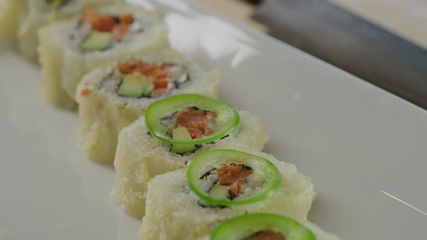 Person garnishes sushi rolls plated on white rectangular tray sitting on wooden surface next to knife