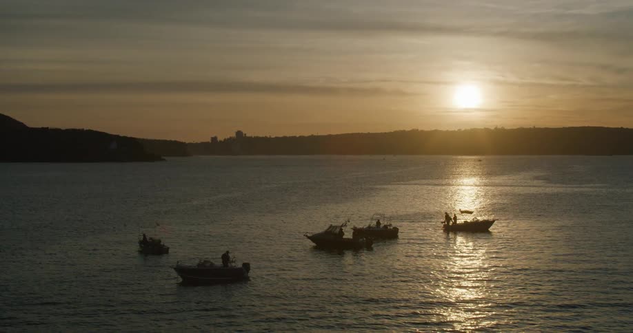 As the sun is set low in the sky several fishers stand at the edge of their boats with their fishing rods
