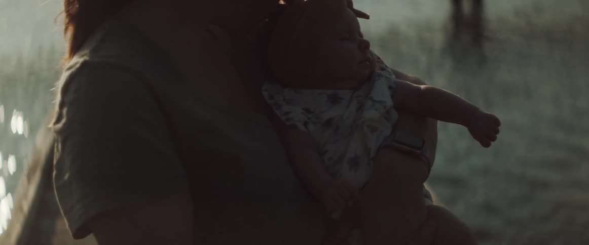 A woman holds her infant daughter close to her chest while standing on a pier on a windy day