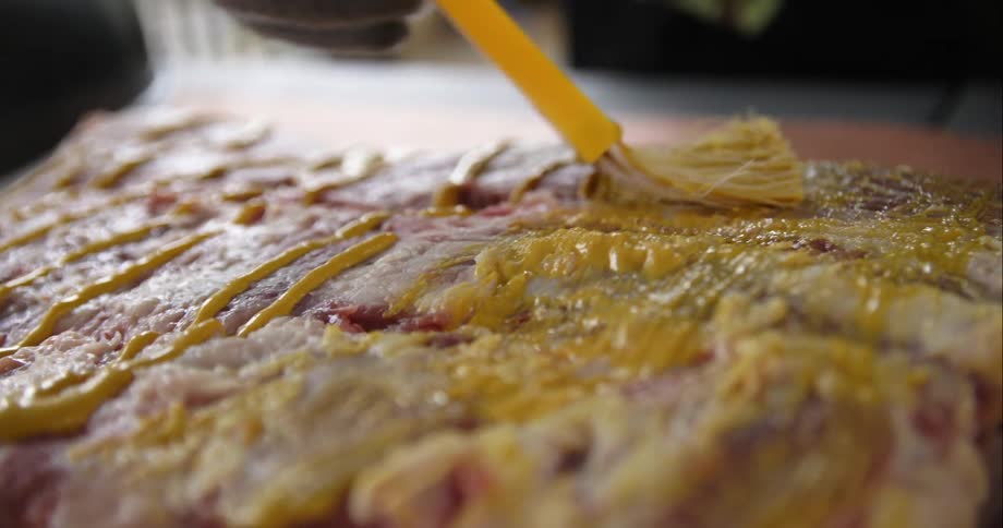 Person wearing gloves spreads mustard onto meat using brush