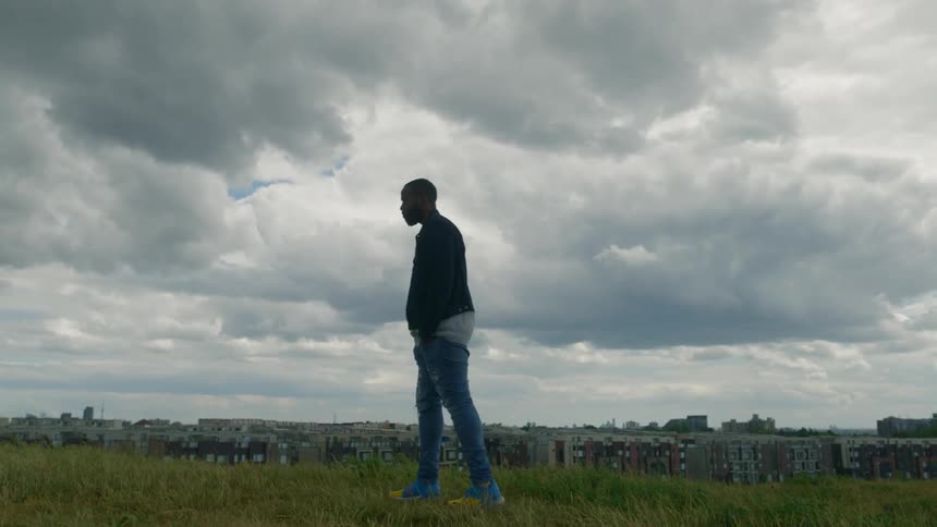 A man standing in a field on the outskirts of a city looks contemplatively into the distance on a cloudy day