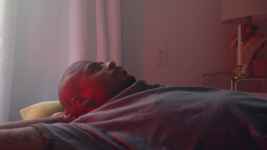 A man lies on a pink bed with his hands behind his head and looks up at the ceiling