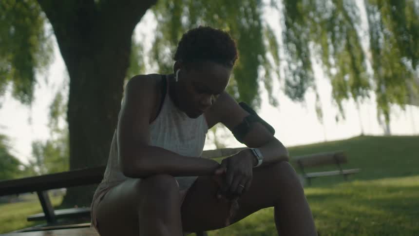 A teenage girl with airpods and her cell phone on her arm sits on a park bench and looks at the ground as she catches her breath