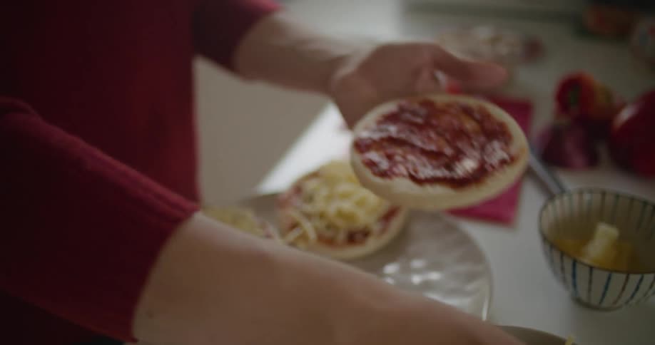 A person in a kitchen adding tomato sauce and cheese to a mini pizza and placing it on a plate on a table