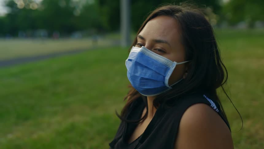 A young woman is wearing a face mask while sitting in the grass at a public park in the evening