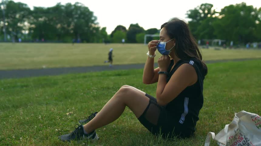 Woman sitting on lawn puts on face mask as man runs by on track with people playing in field in background