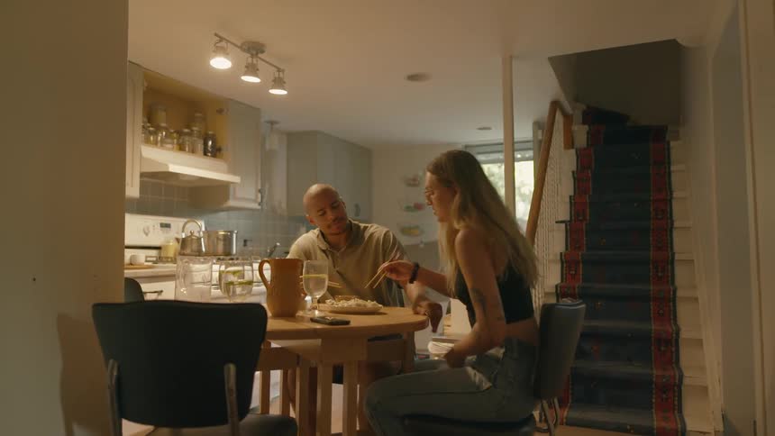 A man and a woman in a room sitting at a table and eating with a wall and a staircase in the background