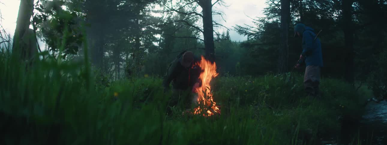 Two men add branches to a campfire in the woods along the river