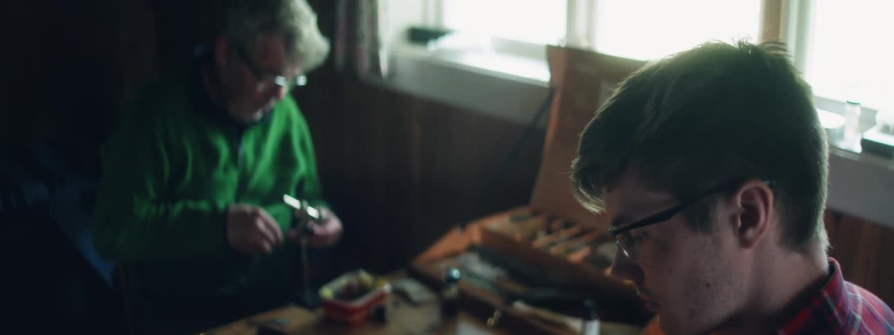 A dad sits at a work table with his son tying a fishing fly