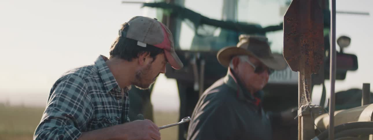 Two men repair machinery standing behind tractor in field
