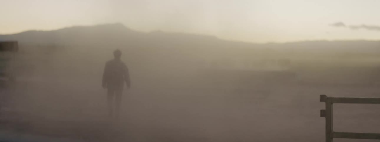 Man wearing checked shirt walks past wooden fence toward mountain disappearing in fog