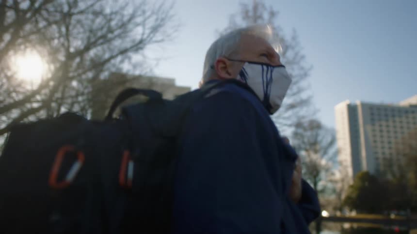 An elderly man walking outside near buildings while wearing a face mask