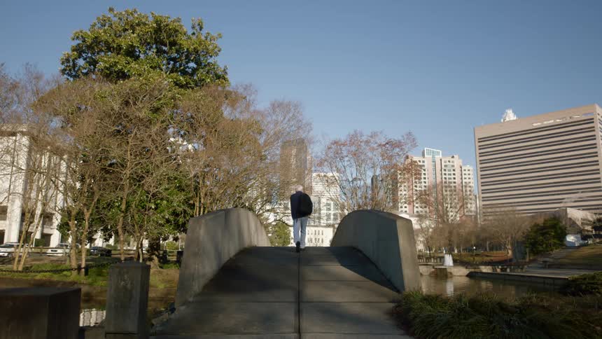 A man carrying a backpack walks across a bridge toward city buildings