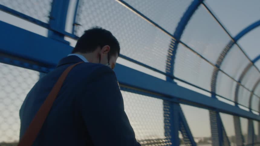 A man holding his phone and carrying a bag walks across a pedestrian bridge on a bright sunny day