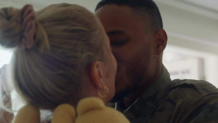 Husband kisses and hugs wife holding a teddy bear in one hand that he give to their baby