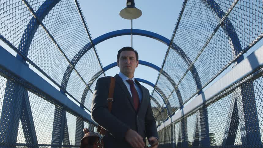 A man with a bag a suit and a tie looking at a smart phone on an overpass with the sky and clouds in the background