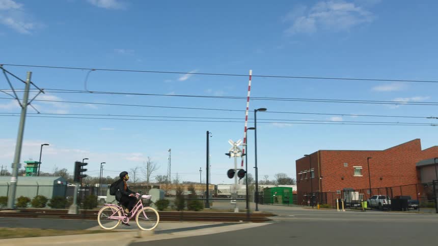 A girl wearing a black helmet is riding a pink bicycle near train tracks and buildings