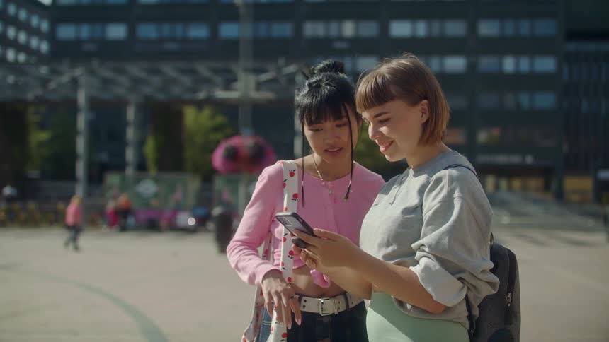 Two girl friends stand in a plaza on a sunny day smiling and laughing as they look at a phone