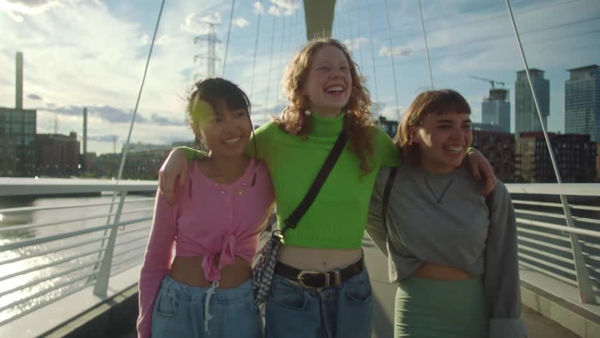Three smiling teen girls crossing a city bridge with their arms around each others shoulders