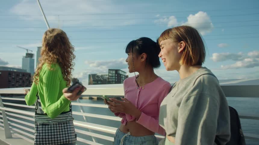 A group of young women standing together on a bridge look at their smartphones together before laughing