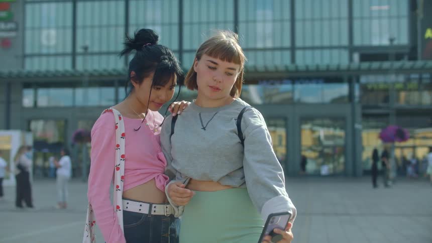 Two women are standing in front of a busy building taking selfies