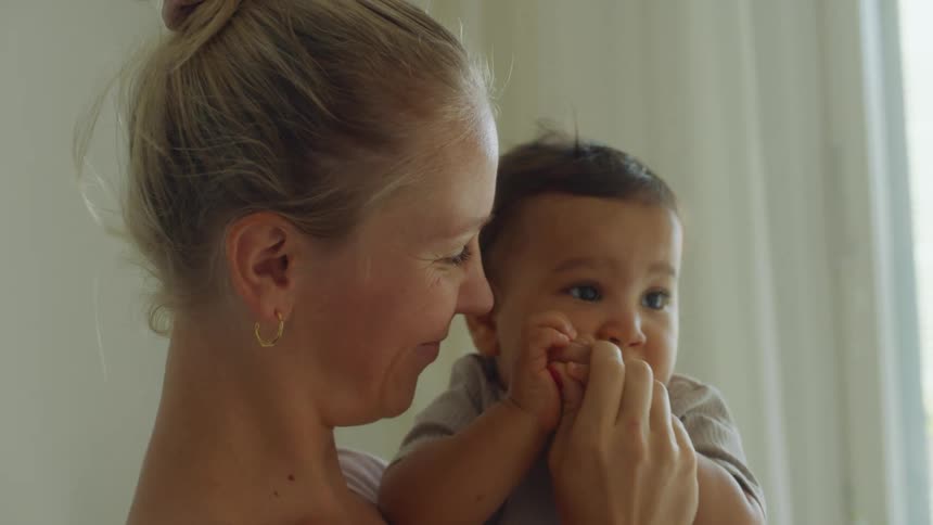 A woman holding and kissing her baby suddenly looks up in surprise as her husband walks over and hugs her
